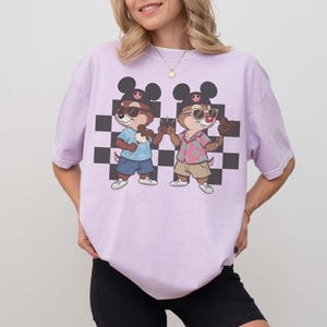 May include: A light purple t-shirt featuring a cartoon graphic of two chipmunks wearing sunglasses and Mickey Mouse hats, holding ice cream cones. The background has a black and white checkered pattern.