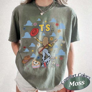 May include: A moss green t-shirt featuring a graphic of a person with long hair playing a guitar, wearing a yellow plaid shirt and cow-print pants. The design includes the text TS, a red hat, blue clouds, flowers, and a stack of blocks labeled TS.