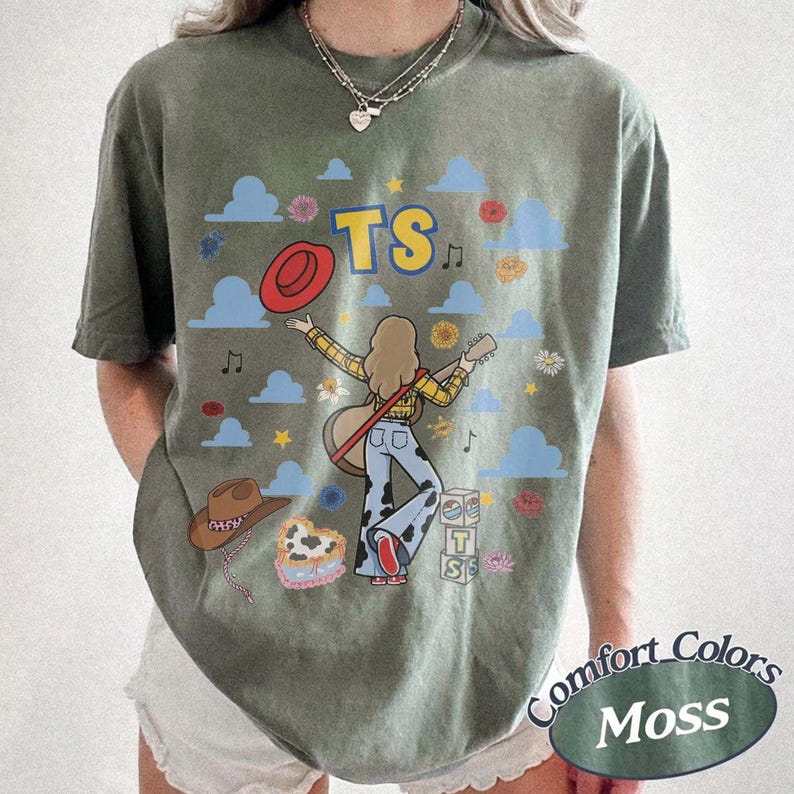 May include: A moss green t-shirt featuring a graphic of a person with long hair playing a guitar, wearing a yellow plaid shirt and cow-print pants. The design includes the text TS, a red hat, blue clouds, flowers, and a stack of blocks labeled TS.