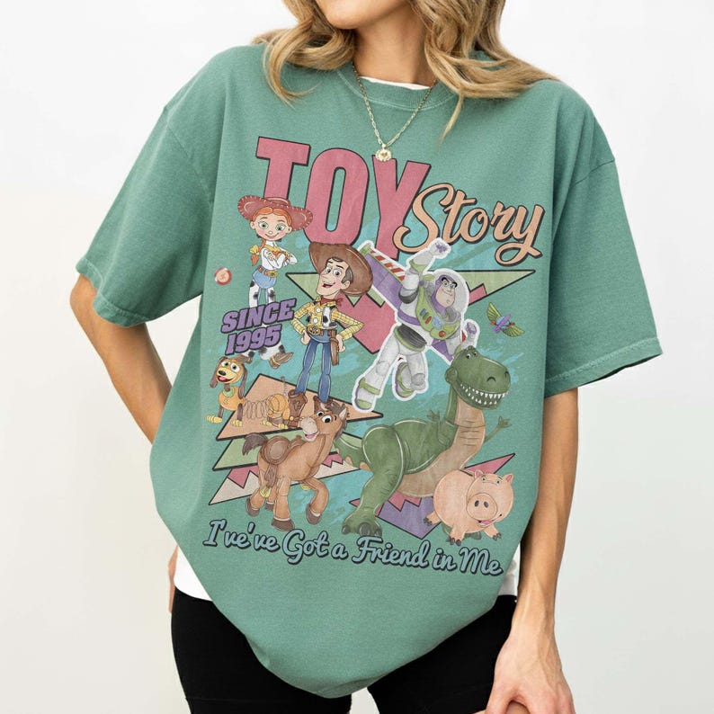 May include: A sage green short-sleeve t-shirt featuring a graphic print of Toy Story characters including Woody, Buzz Lightyear, Jessie, Rex, Slinky Dog, and Hamm. The text reads TOY Story, SINCE 1995, and I've Got a Friend in Me in various fonts.