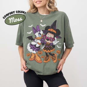 Może przedstawiać: Koszulka w kolorze mchu z grafiką przedstawiającą Daisy Duck i Minnie Mouse w kostiumach na Halloween. Daisy ma na sobie fioletową sukienkę i kokardę, a Minnie kapelusz czarownicy i sukienkę w kropki. Wok&oacute;ł nich znajdują się małe dynie i nietoperze. Tekst głosi COMFORT COLORS Moss.