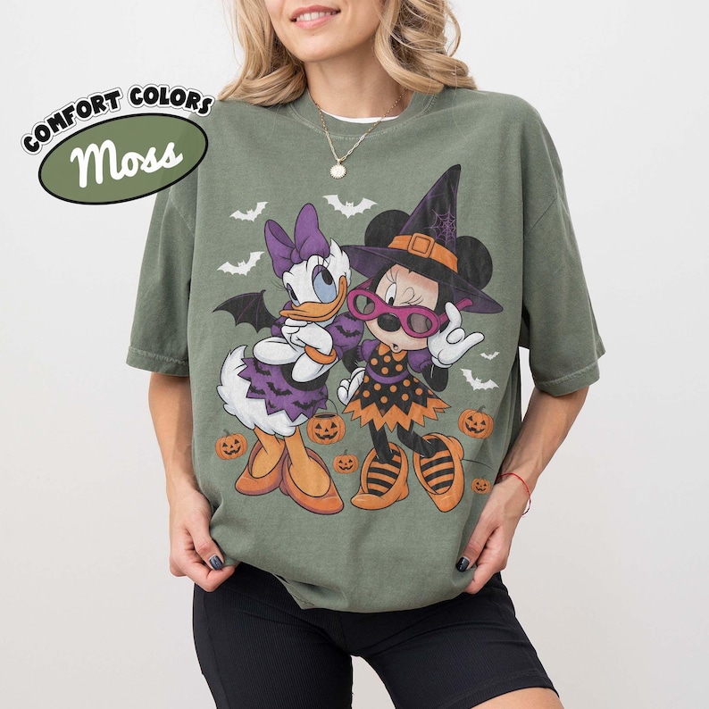 Może przedstawiać: Koszulka w kolorze mchu z grafiką przedstawiającą Daisy Duck i Minnie Mouse w kostiumach na Halloween. Daisy ma na sobie fioletową sukienkę i kokardę, a Minnie kapelusz czarownicy i sukienkę w kropki. Wok&oacute;ł nich znajdują się małe dynie i nietoperze. Tekst głosi COMFORT COLORS Moss.