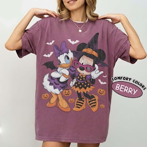 Może przedstawiać: Jagodowy t-shirt z grafiką przedstawiającą Daisy Duck i Minnie Mouse w kostiumach na Halloween. Daisy ma na sobie fioletową sukienkę i kokardę, a Minnie nosi kapelusz czarownicy i okulary. Wz&oacute;r zawiera pomarańczowe dynie oraz nietoperze.