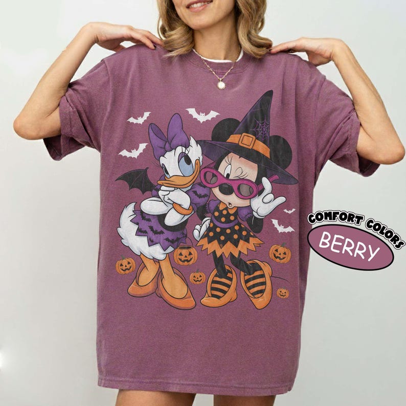 Może przedstawiać: Jagodowy t-shirt z grafiką przedstawiającą Daisy Duck i Minnie Mouse w kostiumach na Halloween. Daisy ma na sobie fioletową sukienkę i kokardę, a Minnie nosi kapelusz czarownicy i okulary. Wz&oacute;r zawiera pomarańczowe dynie oraz nietoperze.