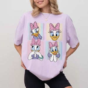 Może przedstawiać: Jasnofioletowa koszulka z czteropanelowym wzorem Kaczki Daisy. Każdy panel przedstawia inną pozę Kaczki Daisy z jej charakterystyczną r&oacute;żową kokardą. Tło każdego panelu ma pastelowe kolory.