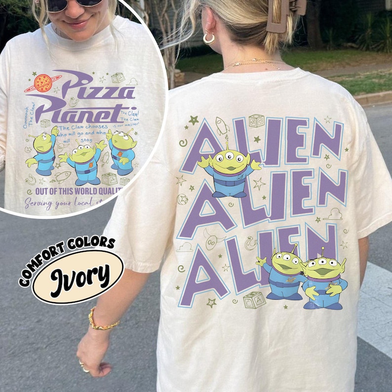 May include: Ivory-colored t-shirt featuring the Pizza Planet logo and alien characters on the front. The back of the shirt displays the word "ALIEN" in purple, along with alien illustrations. The shirt is a comfort color.