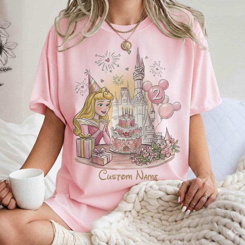 May include: Pale pink t-shirt with a princess illustration, birthday cake, castle, and balloons. The design includes the text "Custom Name" and the number "2" on the balloons. The shirt is a light pink colour.