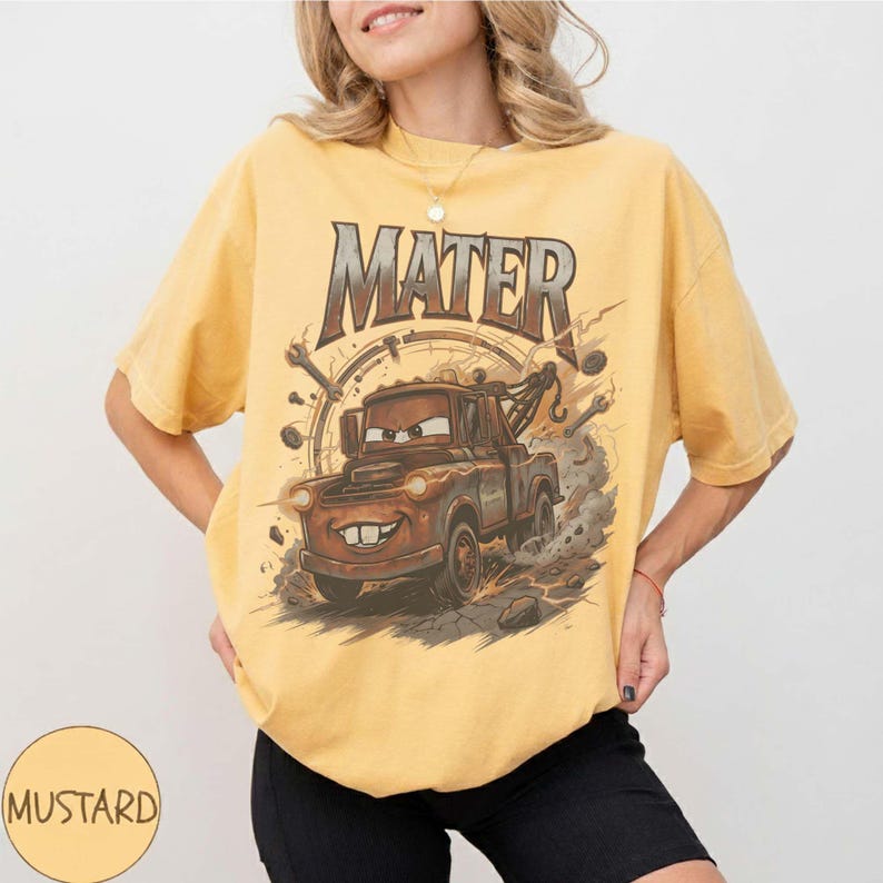 Puede incluir: Camiseta de color mostaza con un gr&aacute;fico de Mate de la pel&iacute;cula Cars. El dise&ntilde;o incluye la palabra "MATER" en una fuente desgastada sobre el cami&oacute;n de remolque de dibujos animados. La camiseta tiene mangas cortas y un corte holgado.
