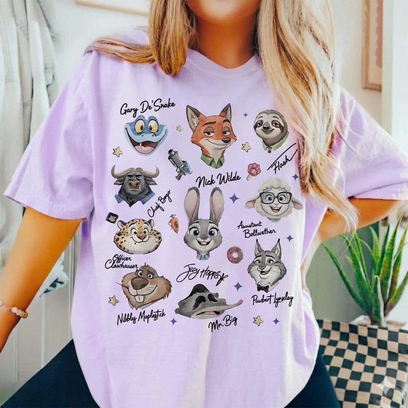 Może przedstawiać: Jasnofioletowa koszulka z postaciami z kresk&oacute;wki Zootopia. Projekt zawiera postacie takie jak Nick Wilde, Judy Hopps i Flash, z ich imionami napisanymi nad lub pod nimi. Koszulka jest elementem odzieży codziennej.