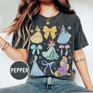 May include: Dark gray t-shirt featuring colorful illustrations of Disney princesses and bows. The shirt has a vintage wash appearance and includes the word "PEPPER" in a circular graphic.