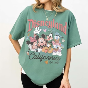 May include: Green t-shirt featuring the Disneyland logo in red script with Mickey Mouse, Minnie Mouse, Donald Duck, Goofy, and Pluto. The shirt also has the text "California" and "Est 1955" in a vintage style.