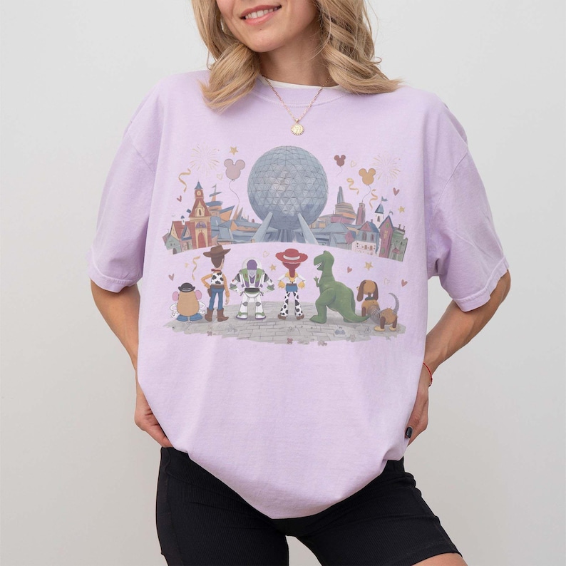 Puede incluir: Camiseta de color lila claro con un gr&aacute;fico colorido de personajes de Toy Story y un parque tem&aacute;tico. El dise&ntilde;o incluye a Woody, Buzz Lightyear y otros personajes. La camiseta tiene cuello redondo y mangas cortas.