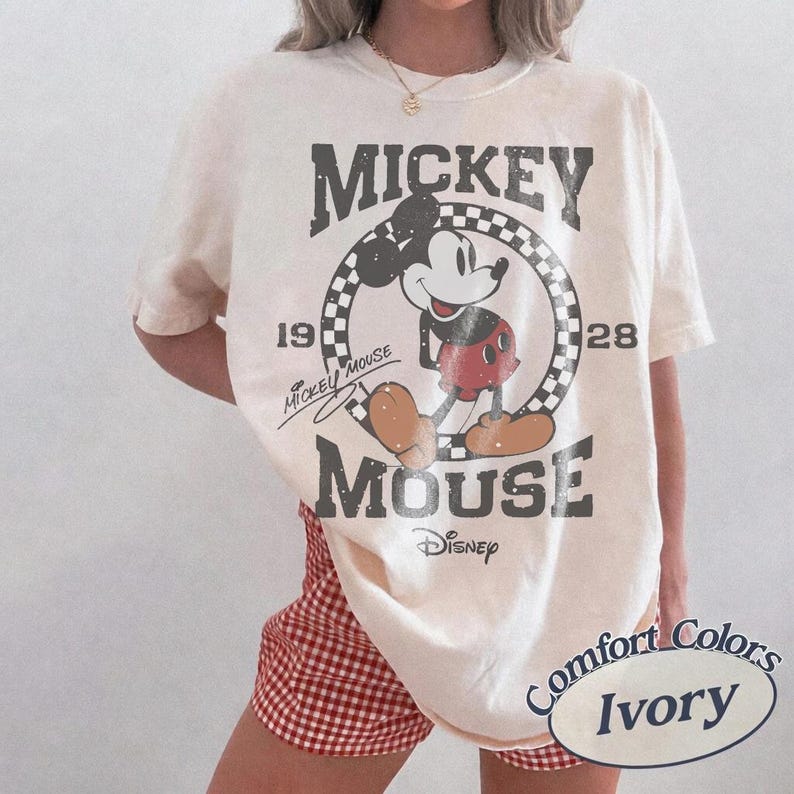 May include: Ivory-colored t-shirt featuring a distressed graphic of Mickey Mouse. The design includes the words "Mickey Mouse" and "Disney" with the years "1928" and "19". The shirt is a Comfort Colors brand.