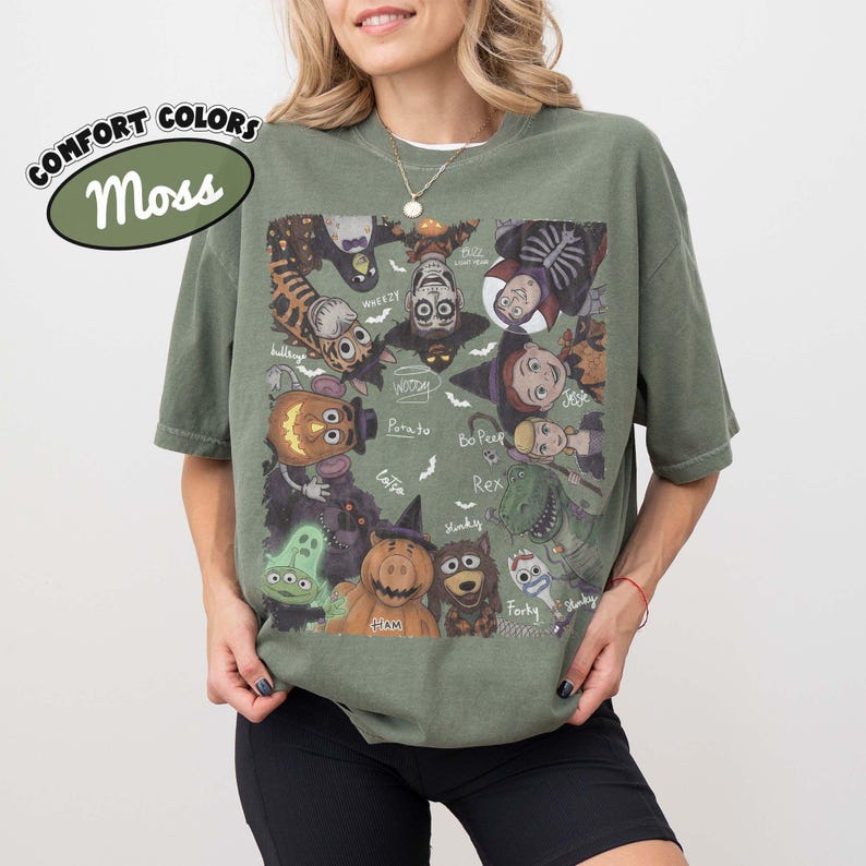 May include: A moss green short-sleeve t-shirt featuring a graphic print of various animated characters dressed in Halloween costumes. The design includes labels for characters like Woody, Potato, Bo Peep, Rex, Forky, Ham, and Wheezy, with small bats.