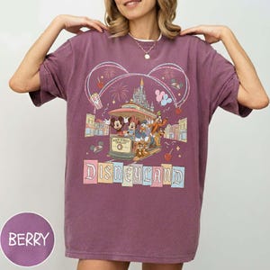 May include: Berry-colored t-shirt featuring a Disneyland graphic. The design includes Mickey Mouse, Minnie Mouse, Donald Duck, and Goofy riding a trolley, with fireworks and the Disneyland logo. The shirt has short sleeves.