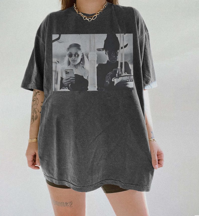May include: Dark gray oversized t-shirt featuring a black and white image of two people. One person is wearing sunglasses and holding a phone, the other is wearing a hat. The shirt has a vintage, worn look.