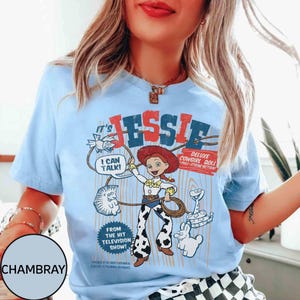 May include: Pale blue t-shirt with a graphic of Jessie from Toy Story. The design includes the text "It's Jessie", "I Can Talk!", and "Deluxe Cowgirl Doll". The shirt is made of a soft material.