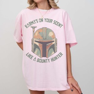 May include: A light pink t-shirt with a Mandalorian helmet graphic in brown, green, and grey. The text "Karma's on your scent like a bounty hunter" is printed above and below the helmet in green.