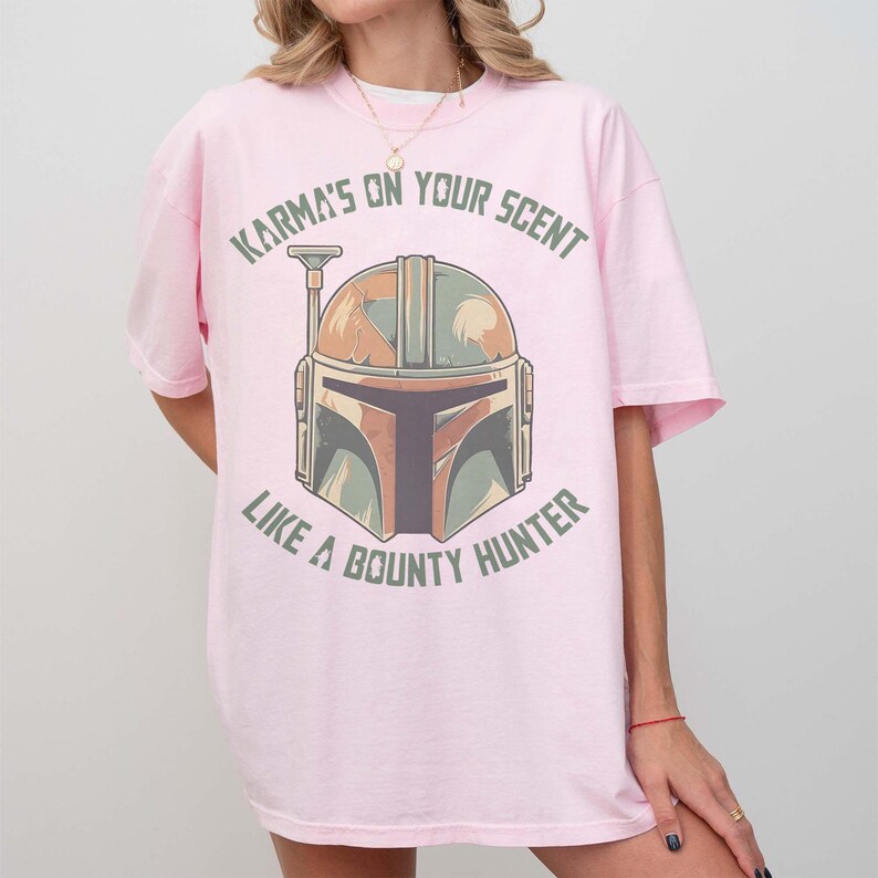 May include: A light pink t-shirt with a Mandalorian helmet graphic in brown, green, and grey. The text "Karma's on your scent like a bounty hunter" is printed above and below the helmet in green.