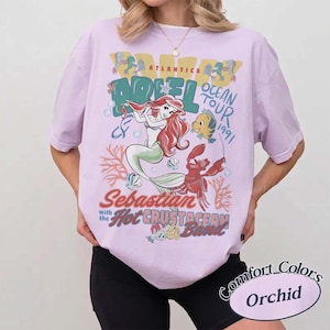 May include: A light purple t-shirt featuring a graphic of Ariel from The Little Mermaid, with the text "Ariel Ocean Tour 1991" and "Sebastian with the Hot Crustacean Band." The shirt is labeled "Comfort Colors Orchid."