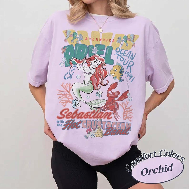 May include: A light purple t-shirt featuring a graphic of Ariel from The Little Mermaid, with the text "Ariel Ocean Tour 1991" and "Sebastian with the Hot Crustacean Band." The shirt is labeled "Comfort Colors Orchid."