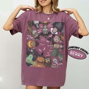 May include: A berry-colored short-sleeve t-shirt featuring a graphic print of various animated characters in Halloween costumes. The design includes labels for characters like Woody, Jessie, Rex, and Forky, surrounded by small white bat illustrations.