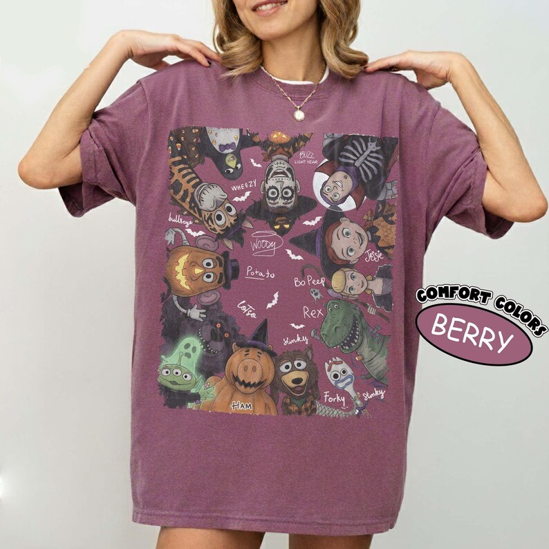 May include: A berry-colored short-sleeve t-shirt featuring a graphic print of various animated characters in Halloween costumes. The design includes labels for characters like Woody, Jessie, Rex, and Forky, surrounded by small white bat illustrations.