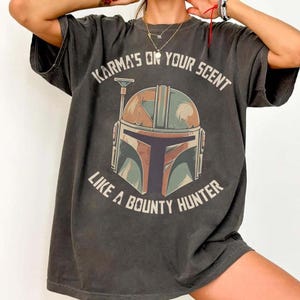May include: Dark grey t-shirt with a Mandalorian helmet graphic and the text "Karma's or your scent like a bounty hunter." The helmet design incorporates brown, green, and blue. The shirt has a vintage, worn appearance.