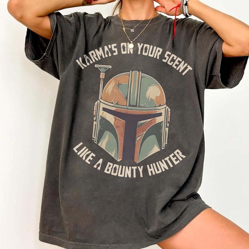 May include: Dark grey t-shirt with a Mandalorian helmet graphic and the text "Karma's or your scent like a bounty hunter." The helmet design incorporates brown, green, and blue. The shirt has a vintage, worn appearance.