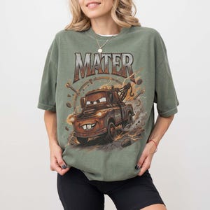 Puede incluir: Camiseta verde oliva con una imagen de dibujos animados de Mater de la pel&iacute;cula Cars. El gr&aacute;fico incluye la palabra "MATER" en una fuente desgastada sobre el personaje. El dise&ntilde;o tiene un aspecto vintage y desgastado.