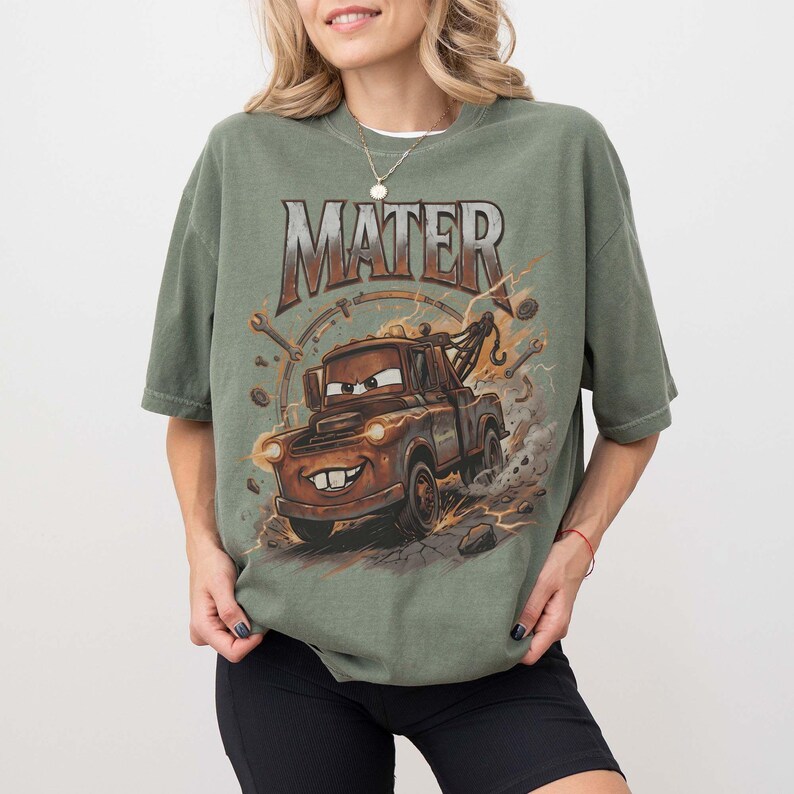 Puede incluir: Camiseta verde oliva con una imagen de dibujos animados de Mater de la pel&iacute;cula Cars. El gr&aacute;fico incluye la palabra "MATER" en una fuente desgastada sobre el personaje. El dise&ntilde;o tiene un aspecto vintage y desgastado.