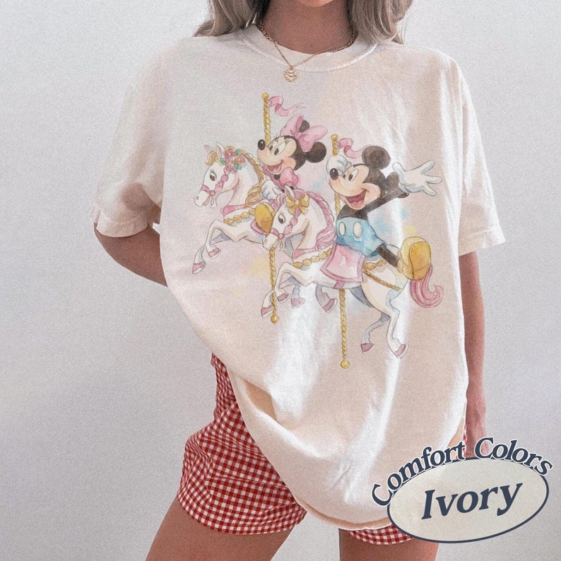 Op de afbeelding: Ivoor kleurig T-shirt met een carrousel sc&egrave;ne met Mickey en Minnie Mouse op witte paarden. De afbeelding bevat pastelkleuren en gouden accenten. Het shirt is van het merk Comfort Colors.