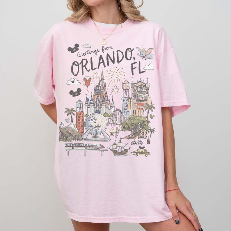 K&ouml;nnte beinhalten: Hellrosa T-Shirt mit farbenfroher Grafik von Wahrzeichen von Orlando, Florida, darunter ein Schloss, Feuerwerk und der Text "Greetings from Orlando, FL". Das Shirt hat kurze &Auml;rmel und eine lockere Passform.
