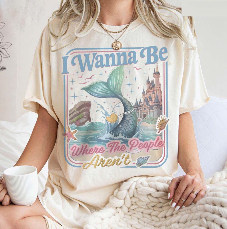 May include: Cream-colored t-shirt with a mermaid graphic and the text "I Wanna Be Where The People Aren't." The design features a mermaid tail, castle, and ocean elements in blue, pink, and yellow. The shirt has short sleeves.