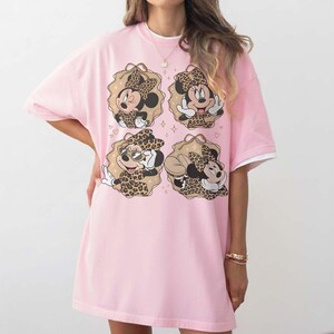 May include: Pink t-shirt dress featuring four Minnie Mouse illustrations in leopard print outfits. The illustrations are set within decorative frames. The dress has short sleeves with white trim and a round neckline.