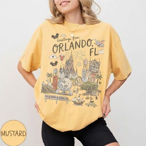 K&ouml;nnte beinhalten: Senfgelbes T-Shirt mit einer Grafik von Orlando, Florida, mit einem Schloss, Feuerwerk und einer Einschienenbahn. Der Text "Greetings from Orlando, FL" ist &uuml;ber der Illustration aufgedruckt. Das Shirt ist kurz&auml;rmlig und hat einen Rundhalsausschnitt.