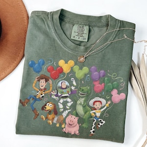 May include: An olive green short-sleeve t-shirt featuring a graphic print of five animated characters from Toy Story, including Woody, Buzz Lightyear, Rex, Jessie, and Slinky Dog, surrounded by colorful Mickey Mouse-shaped balloons and confetti.