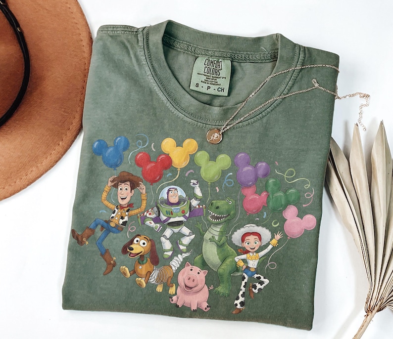 May include: An olive green short-sleeve t-shirt featuring a graphic print of five animated characters from Toy Story, including Woody, Buzz Lightyear, Rex, Jessie, and Slinky Dog, surrounded by colorful Mickey Mouse-shaped balloons and confetti.