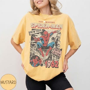 K&ouml;nnte beinhalten: Senfgelbes T-Shirt mit einem Vintage-Spider-Man-Motiv. Das Design enth&auml;lt den Text "THE AMAZING SPIDER-MAN NEW YORK" und "1962". Der Superheld ist in seinem klassischen rot-blauen Anzug zu sehen.