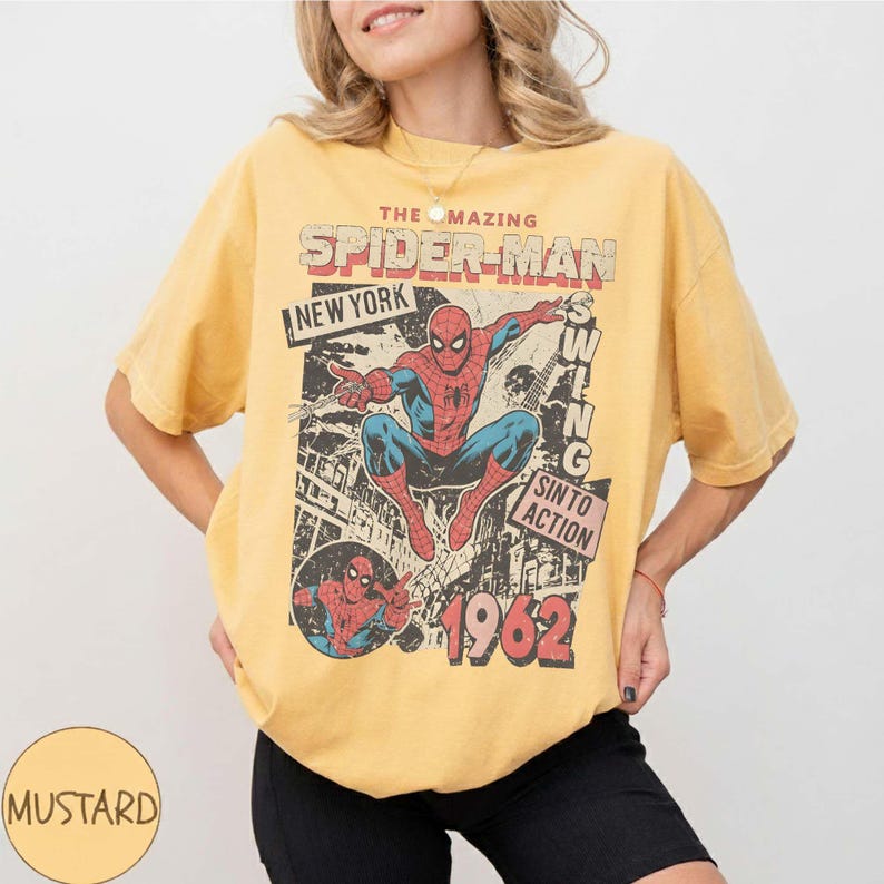K&ouml;nnte beinhalten: Senfgelbes T-Shirt mit einem Vintage-Spider-Man-Motiv. Das Design enth&auml;lt den Text "THE AMAZING SPIDER-MAN NEW YORK" und "1962". Der Superheld ist in seinem klassischen rot-blauen Anzug zu sehen.