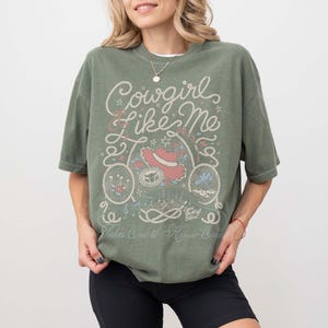 May include: An olive green short-sleeve t-shirt featuring a graphic print with the text Cowgirl Like Me, Eyes full of Stars, and Takes One to Know One. The design includes illustrations of a cowboy hat, rope, horseshoe, and musical notes.