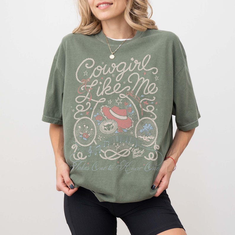 May include: An olive green short-sleeve t-shirt featuring a graphic print with the text Cowgirl Like Me, Eyes full of Stars, and Takes One to Know One. The design includes illustrations of a cowboy hat, rope, horseshoe, and musical notes.