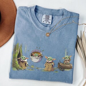 May include: A blue crew neck t-shirt featuring a graphic print of four small green creatures with large ears wearing tan robes. One creature is in a tree hollow, one in a floating pod, one standing, and one sitting on a log. The label reads Comfort Colors.