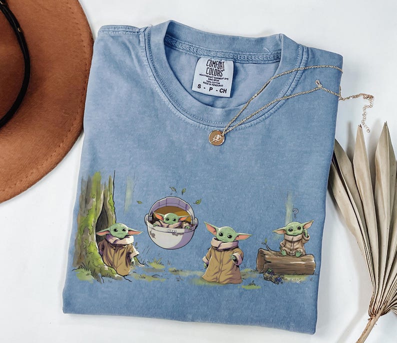May include: A blue crew neck t-shirt featuring a graphic print of four small green creatures with large ears wearing tan robes. One creature is in a tree hollow, one in a floating pod, one standing, and one sitting on a log. The label reads Comfort Colors.