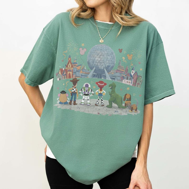 Puede incluir: Camiseta verde con un dise&ntilde;o colorido de personajes de Disney, incluyendo a Woody y Buzz Lightyear, frente a una esfera futurista y edificios. El dise&ntilde;o presenta una variedad de colores y tiene un corte relajado. La camiseta tiene mangas cortas.