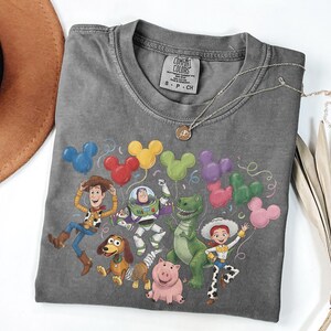 May include: A charcoal gray crew neck t-shirt featuring a graphic print of Toy Story characters including Woody, Buzz Lightyear, Rex, Jessie, Slinky Dog, and Hamm surrounded by colorful Mickey Mouse-shaped balloons. The shirt has a Comfort Colors label.