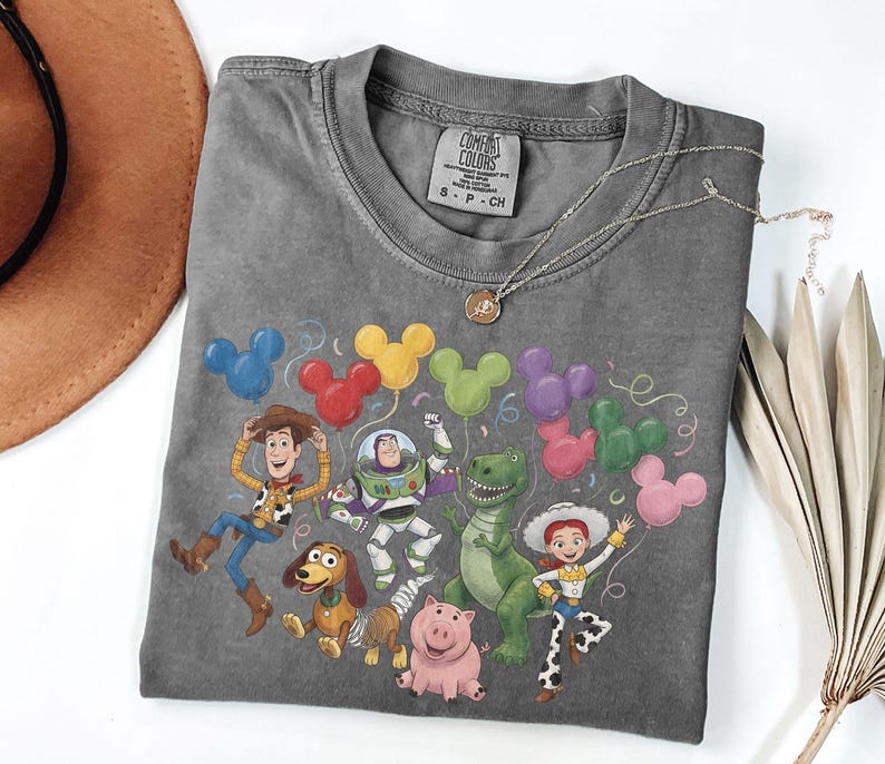 May include: A charcoal gray crew neck t-shirt featuring a graphic print of Toy Story characters including Woody, Buzz Lightyear, Rex, Jessie, Slinky Dog, and Hamm surrounded by colorful Mickey Mouse-shaped balloons. The shirt has a Comfort Colors label.