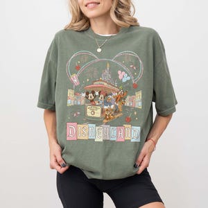 May include: Olive green t-shirt featuring a Disneyland graphic. The design includes Mickey and Minnie Mouse, Donald Duck, Goofy, and Chip and Dale riding a trolley. The words "Main Street Trolley" and "Disneyland" are also visible.