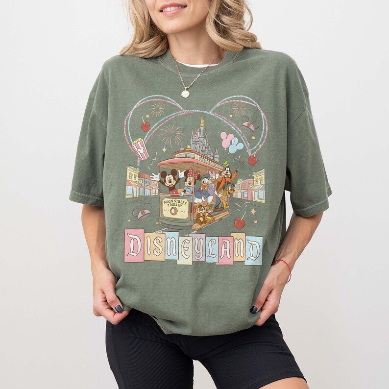 May include: Olive green t-shirt featuring a Disneyland graphic. The design includes Mickey and Minnie Mouse, Donald Duck, Goofy, and Chip and Dale riding a trolley. The words "Main Street Trolley" and "Disneyland" are also visible.