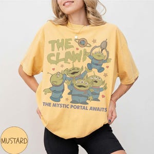 May include: Mustard yellow t-shirt featuring a graphic of green aliens with three eyes, wearing blue uniforms, and the text "THE CLAW!" and "THE MYSTIC PORTAL AWAITS". The design includes stars and a planet.